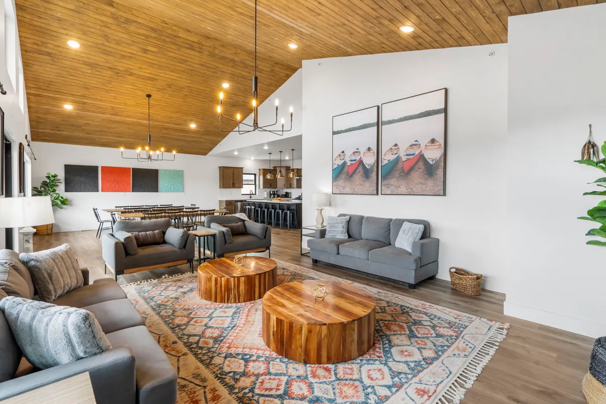 The image showcases a warm, inviting living space with a striking wooden ceiling, plush comfortable sofas arranged for conversation, oversized coffee tables perfect for gathering, and vibrant artwork that adds a pop of color and personality. The room is spacious and clearly designed for families and groups to relax, play games, or enjoy memorable meals together.