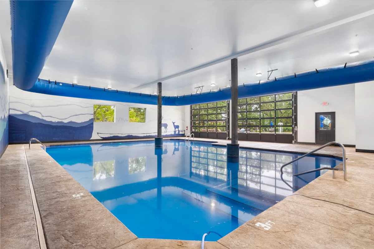 This interior shot showcases a bright, inviting indoor swimming pool with large glass doors letting in natural light and a decorative mural adding artistic flair. The pool is ideal for swimming, relaxing, and socializing year-round.