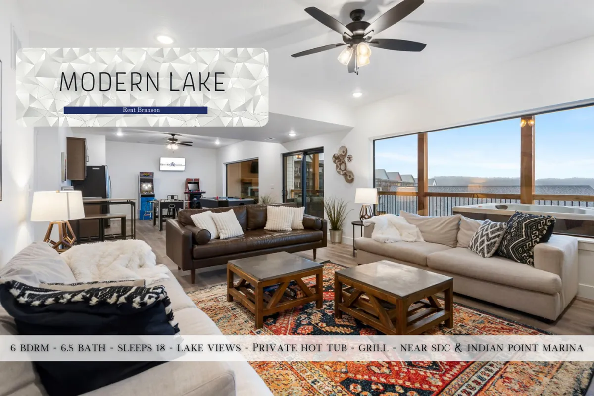 This image showcases a spacious living room with plush, ample seating arranged around a stylish area rug. Large windows dominate one wall, offering uninterrupted lake views and bathing the room in natural light. The arrangement is perfect for movie nights or relaxed conversations, with a modern ceiling fan overhead enhancing comfort and ambiance.