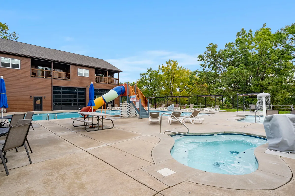 A vibrant scene showing a large outdoor swimming pool area equipped with a winding water slide, hot tubs, lounge chairs, and the building facade in the background. The area is bustling with opportunities for relaxation and play, suitable for families and groups.