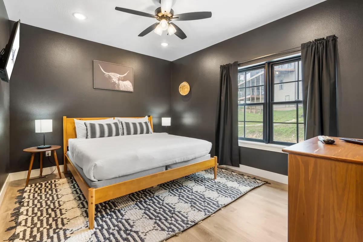 A spacious bedroom with a comfortable bed adorned with plush bedding, flanked by nightstands and a window letting in natural sunlight. Decorative artwork and a cozy rug set a relaxing, inviting atmosphere, perfect for unwinding after a busy day.