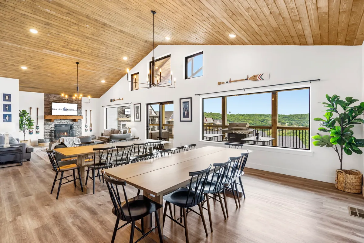 This interior photo captures an inviting dining area seamlessly connected to a cozy living space. The large dining table is surrounded by comfortable seating and positioned near expansive windows, which let in ample natural light. A striking chandelier hangs above, and a prominent indoor fireplace anchors the living area, where plush sofas invite relaxation. The space is styled for both festive celebrations and casual mornings, with lake views visible through the windows.