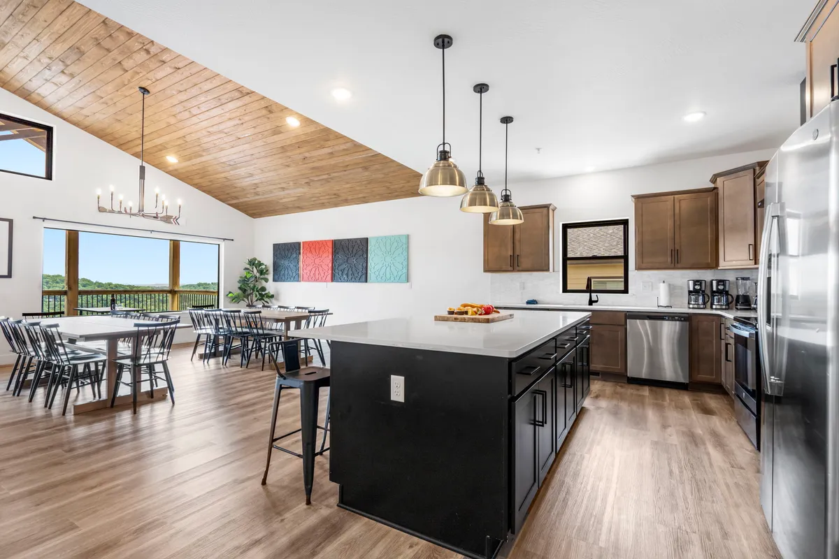 This image presents a welcoming open-concept kitchen and dining area. A large kitchen island serves as a focal point, surrounded by stylish bar stools and complemented by colorful wall art. The dining table is set beside expansive windows, allowing natural light to flood the space and highlighting scenic lake views beyond. The modern appliances and generous counter space are visible, creating an inviting atmosphere for group meal preparations and shared dining experiences.