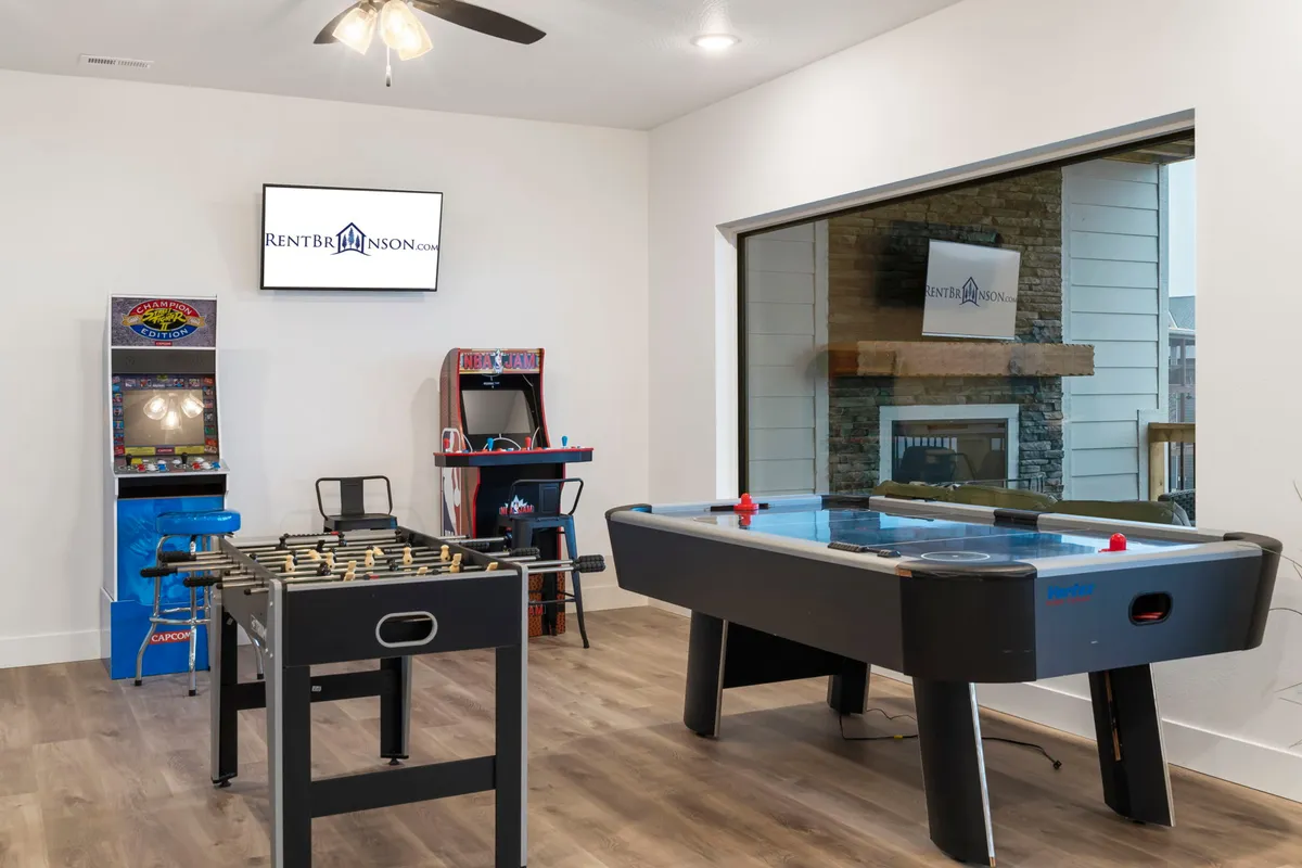 This image captures the heart of the lower level entertainment area, featuring a foosball table, air hockey table, arcade game machines, and a television. The setup creates an inviting atmosphere for group play and friendly competition, with ample space for socializing and laughter.