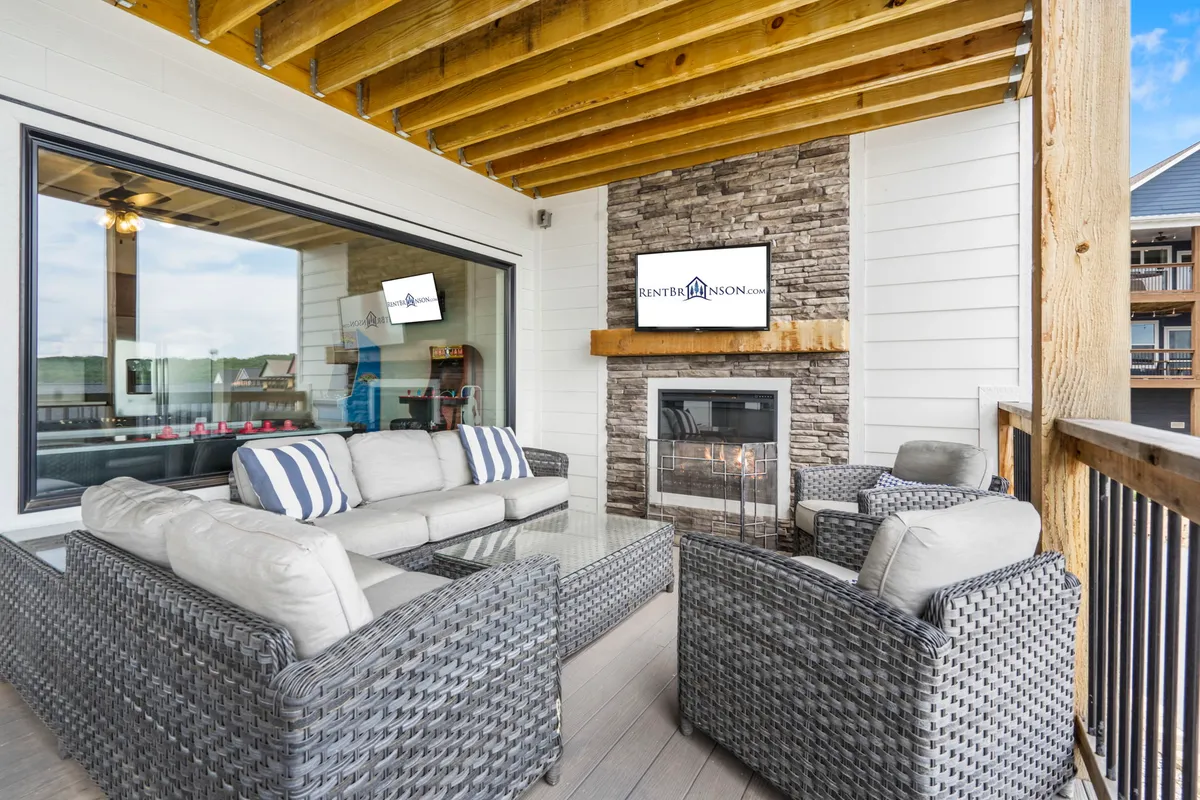 An inviting outdoor seating area complete with a built-in fireplace and television, surrounded by lush landscaping. Comfortable chairs face the fireplace, making it a perfect spot to gather and enjoy outdoor entertainment regardless of the weather.
